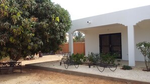 A covered outdoor area with a white building, a large tree, and a metal bench. - LITTLE PIECE OF HEAVEN (SOMONE)
