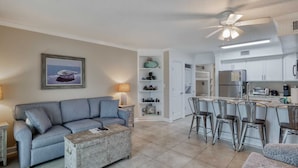 A living room with a grey sofa, a coffee table, and a painting of a boat. There is a kitchen with white cabinets, a refrigerator, and a bar area with stools. - Beachfront Family Condo w/ Balcony Views, Hot Tub, Pool & Great Rates (Gulf Shores)