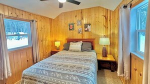 Quarto com painéis de madeira, cama, duas mesas de cabeceira, ventoinha no teto e janela com vista para árvores.