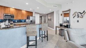 A modern kitchen with wooden cabinets, a microwave, and a stainless steel refrigerator. There are two bar stools and a grey sofa in the living area. - Beautiful Upscale 6th floor Condo (Cape Coral)