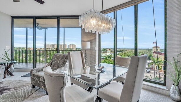 A spacious living room with a glass table, comfortable seating, a decorative chandelier, and a large window offering a city view. - Beautiful Upscale 6th floor Condo (Cape Coral)