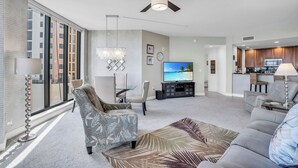A modern living room with a large TV, a ceiling fan, and a dining area with a table and chairs. - Beautiful Upscale 6th floor Condo (Cape Coral)
