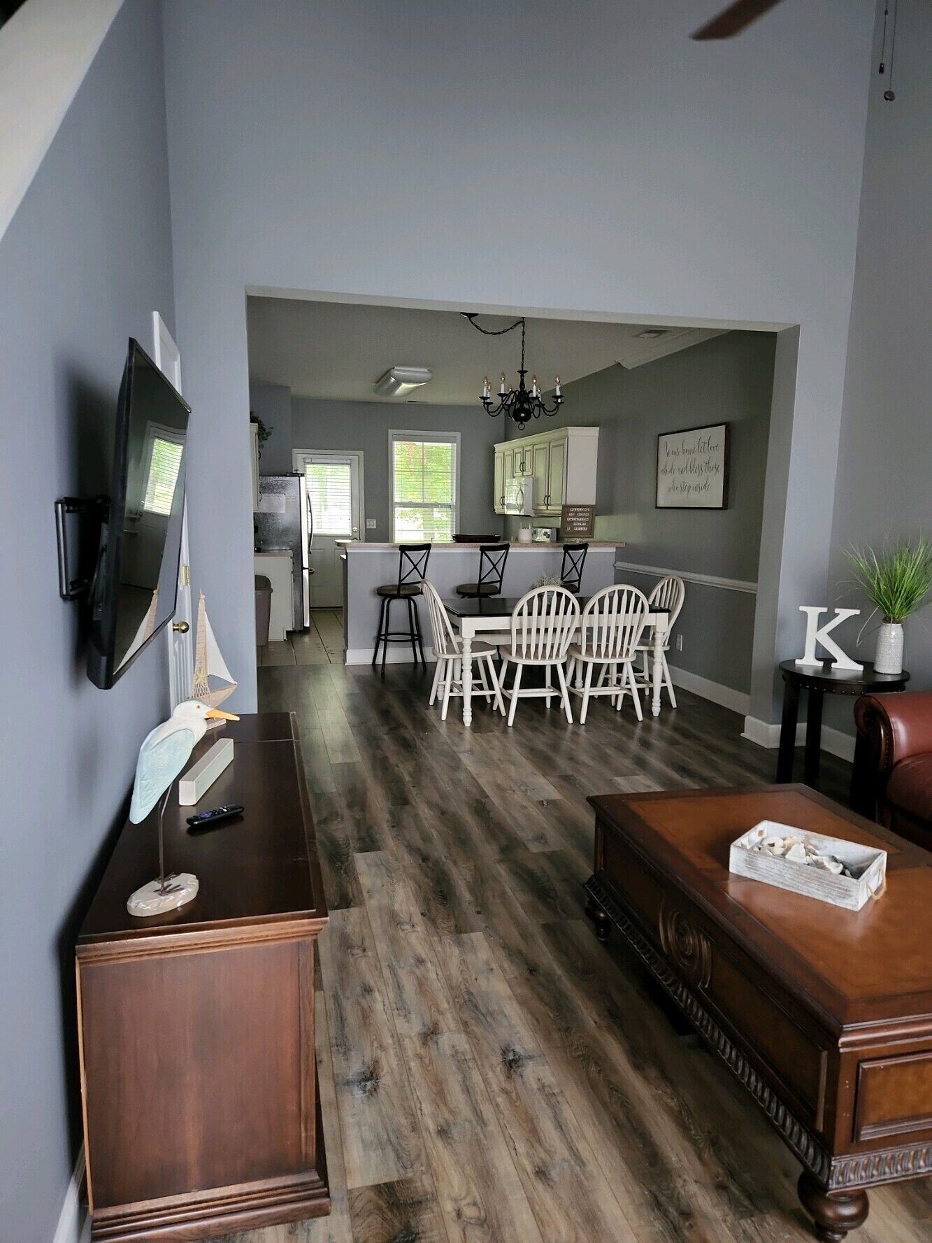 A spacious living room with a dark wood floor, a wooden coffee table, and a TV on a stand. There is a dining area with white chairs and a kitchen in the background.