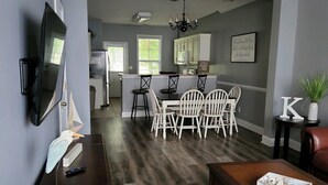 A spacious living room with a dark wood floor, a wooden coffee table, and a TV on a stand. There is a dining area with white chairs and a kitchen in the background. - Come spend a few days by  Barefoot Landing available July 10-14 (North Myrtle Beach)