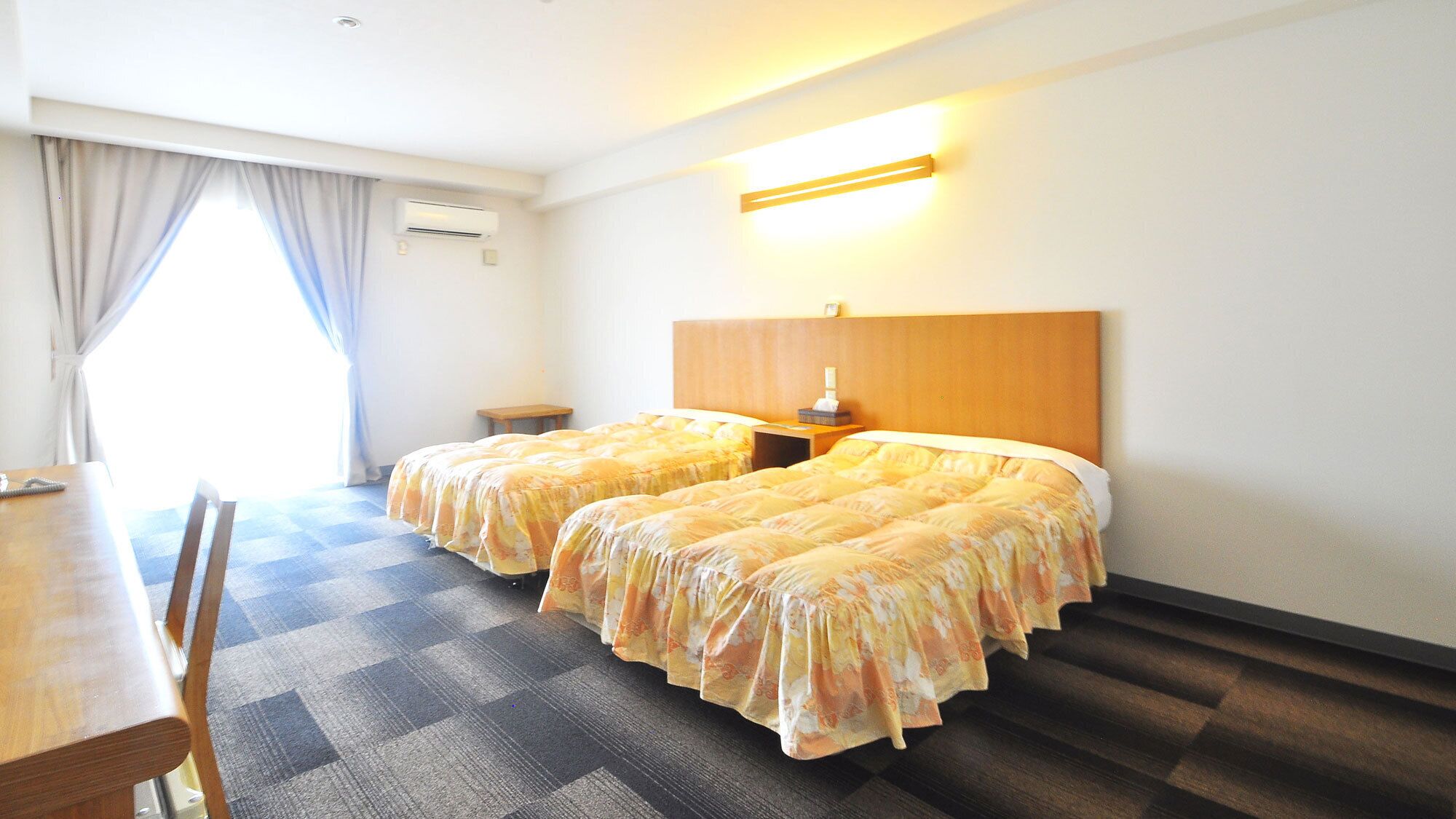 Une chambre d’hôtel avec deux lits, une tête de lit en bois, une table de chevet avec une lampe, une petite table avec une chaise et une fenêtre avec des rideaux.