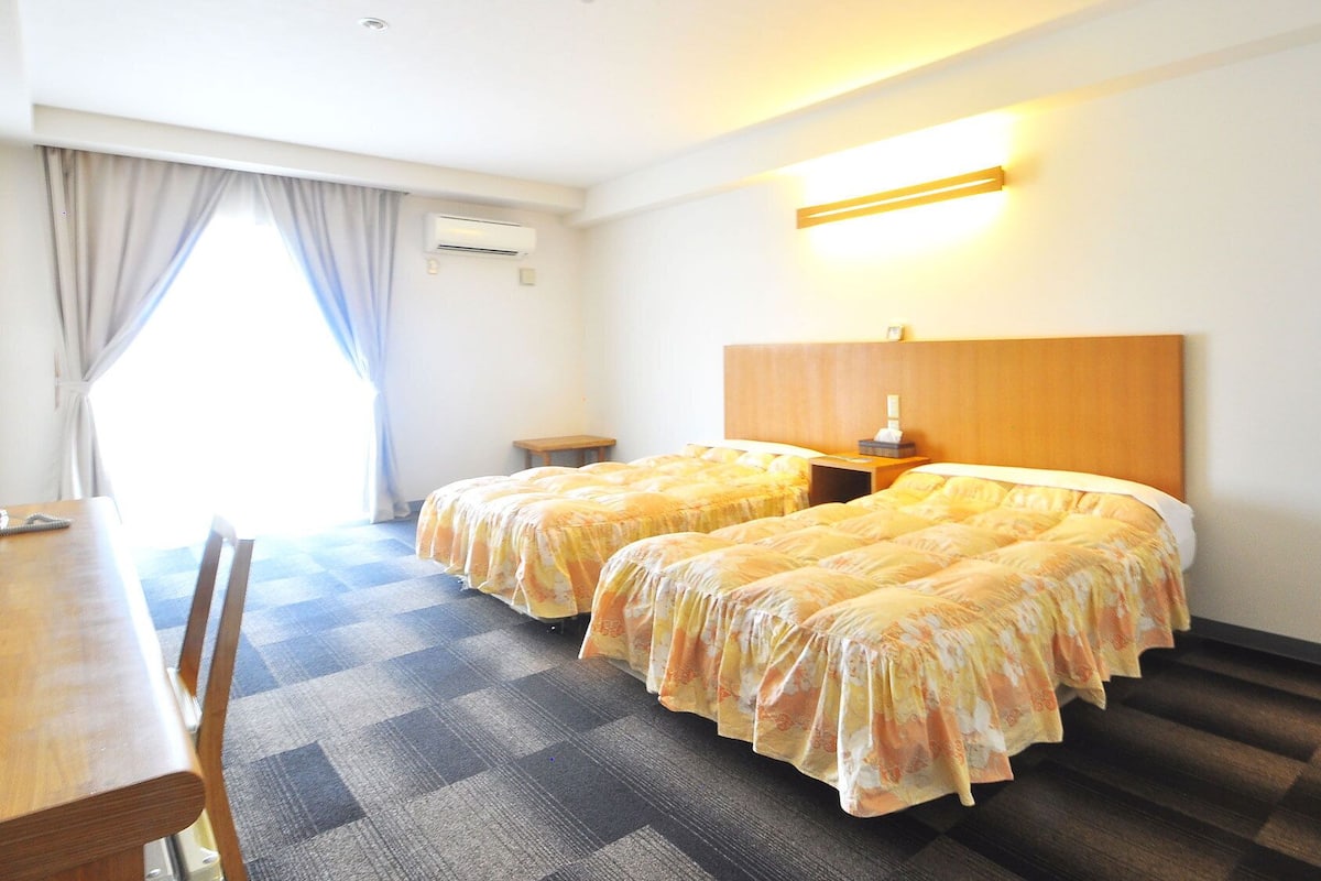 Ein Hotelzimmer mit zwei Betten, einem hölzernen Kopfteil, einem Nachttisch mit Lampe, einem kleinen Tisch mit Stuhl und einem Fenster mit Vorhängen.