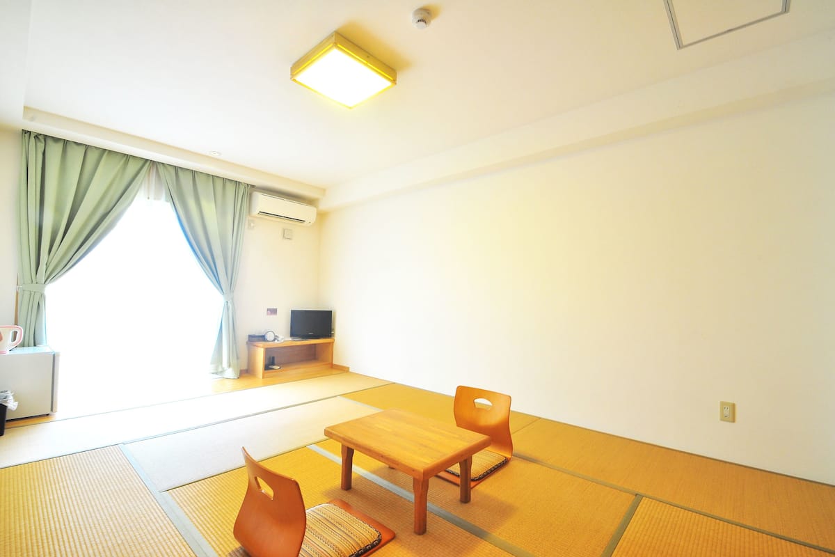 Ein Zimmer mit Tatami-Matten, einem Holztisch, einem Stuhl, einem Fernseher und einem Vorhangfenster.