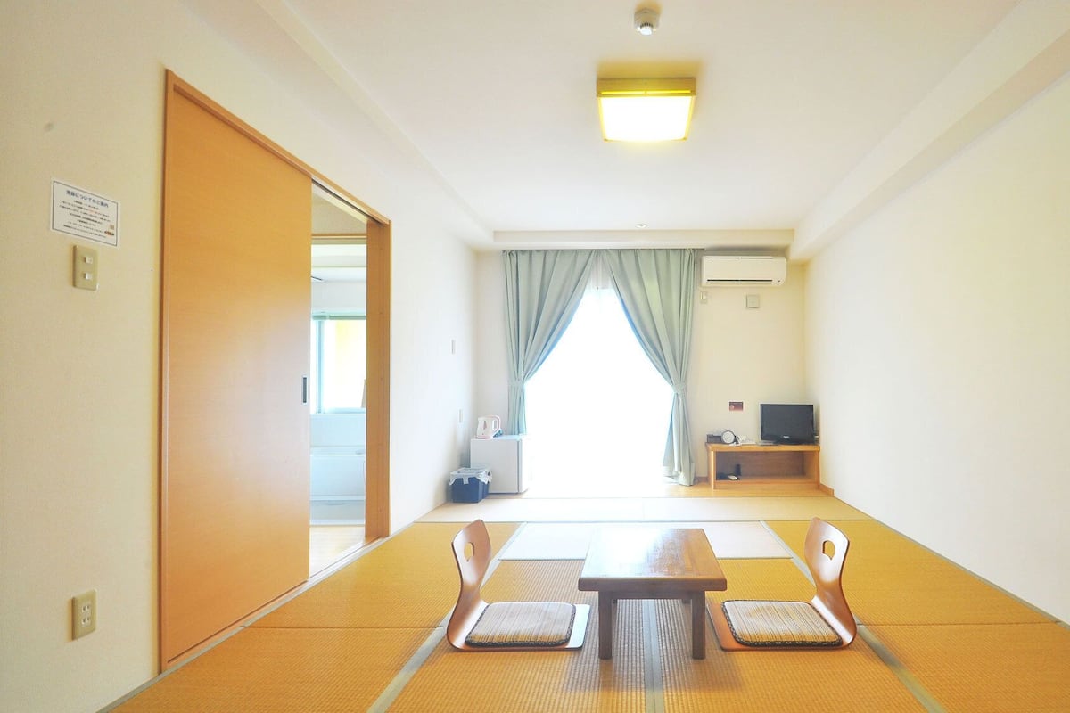 Ein Zimmer mit Tatami-Matten, einem kleinen Tisch, Stühlen, einem Fernseher und einer Schiebetür.