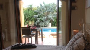 Une chambre d’hôtel avec un lit, une table sur laquelle est posé un chandelier, une chaise, et une vue sur une piscine et la verdure environnante.