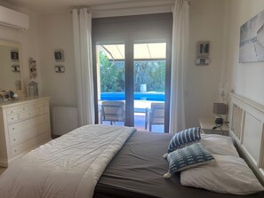 A bedroom with a bed, a dresser, a radiator, a window with curtains, a wall-mounted lamp, and a view of an outdoor pool area.