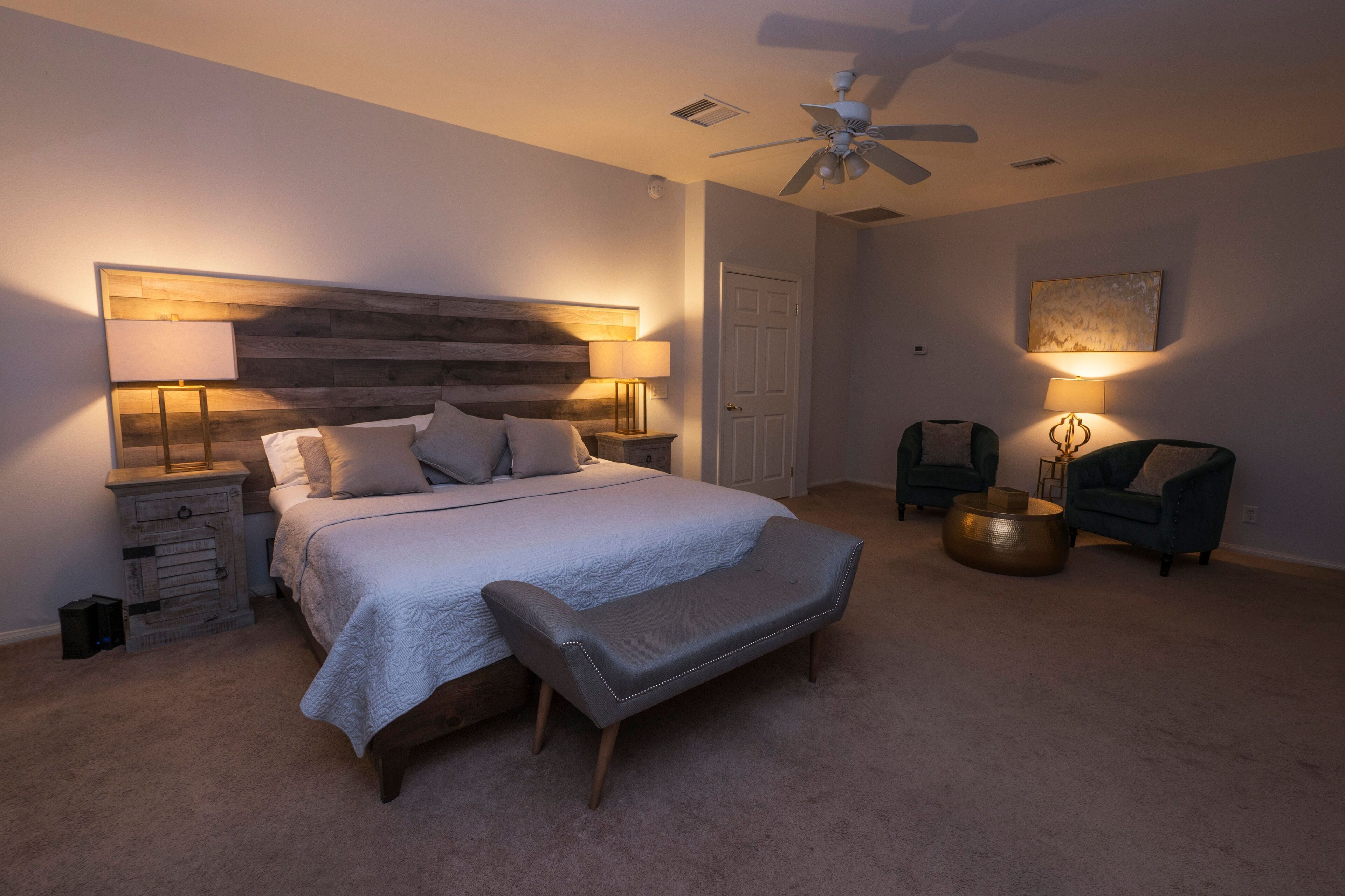 A bedroom with a bed, bedside tables, a bench, a chair, a ceiling fan, and a door.