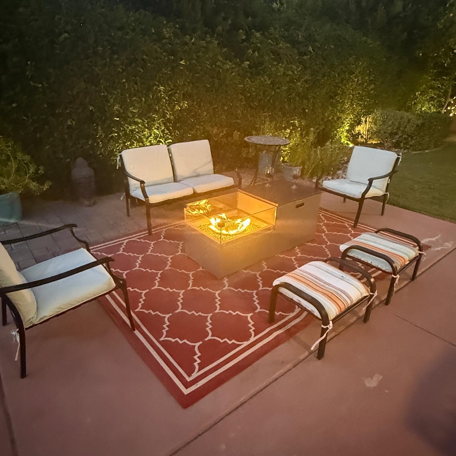 Eine Terrasse im Freien mit einem Feuerkorb mit Glasplatte, umgeben von vier weißen Polsterstühlen und zwei gestreiften Polsterbänken.