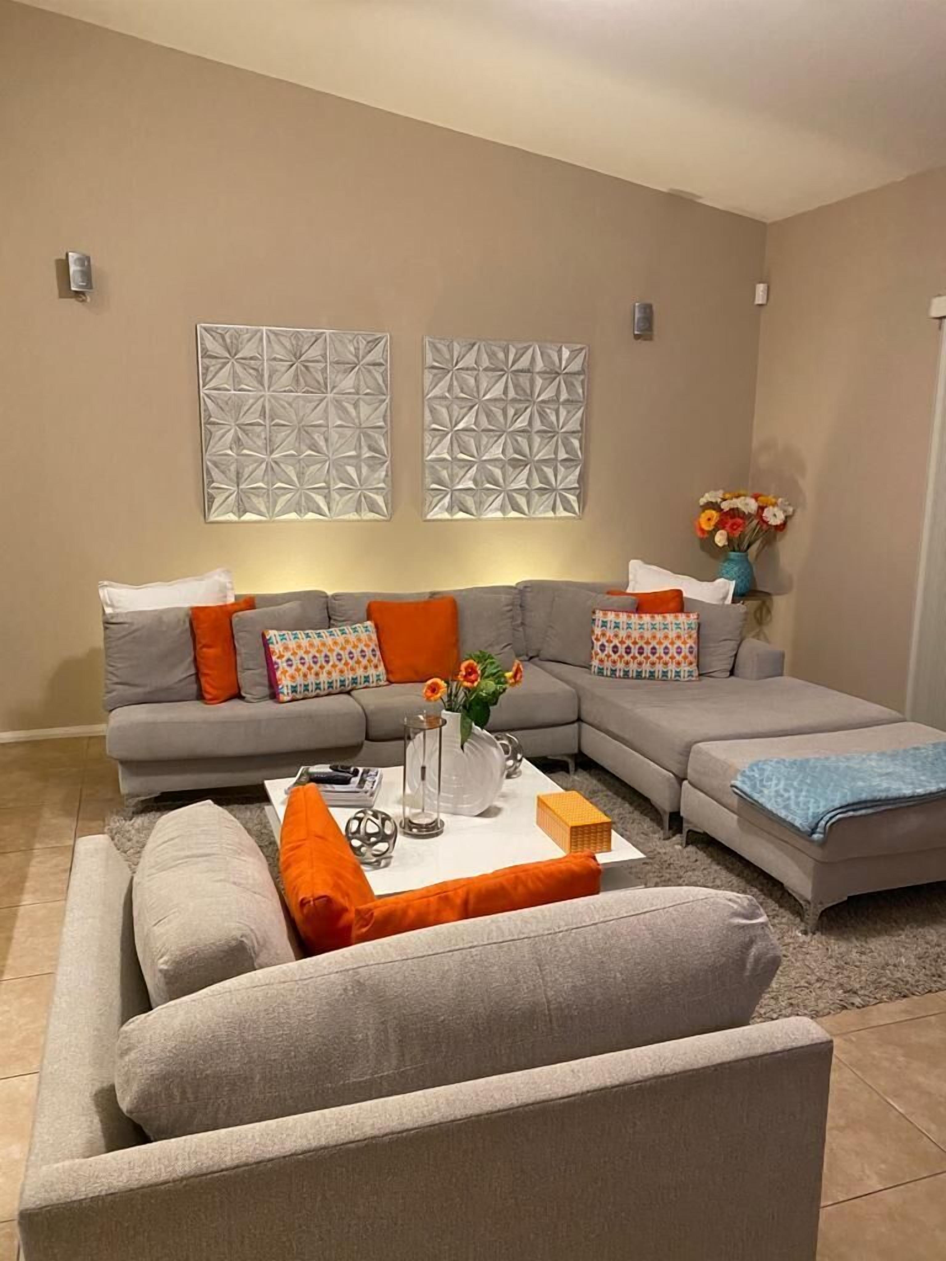 A living room with a grey sectional sofa, orange throw pillows, a coffee table with a vase of flowers, and two wall-mounted geometric art pieces.
