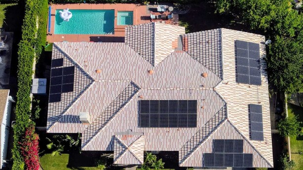 Luchtbeeld van een huis met een uniek geometrisch dakontwerp, zonnepanelen, een zwembad en een verzorgde tuin.