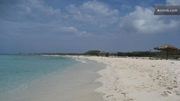 A beach with clear turquoise water, white sand, and thatched umbrellas.