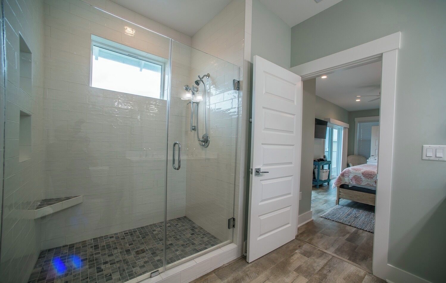 A modern bathroom with a glass-enclosed shower, a white door, and a visible bedroom with a bed and a window.