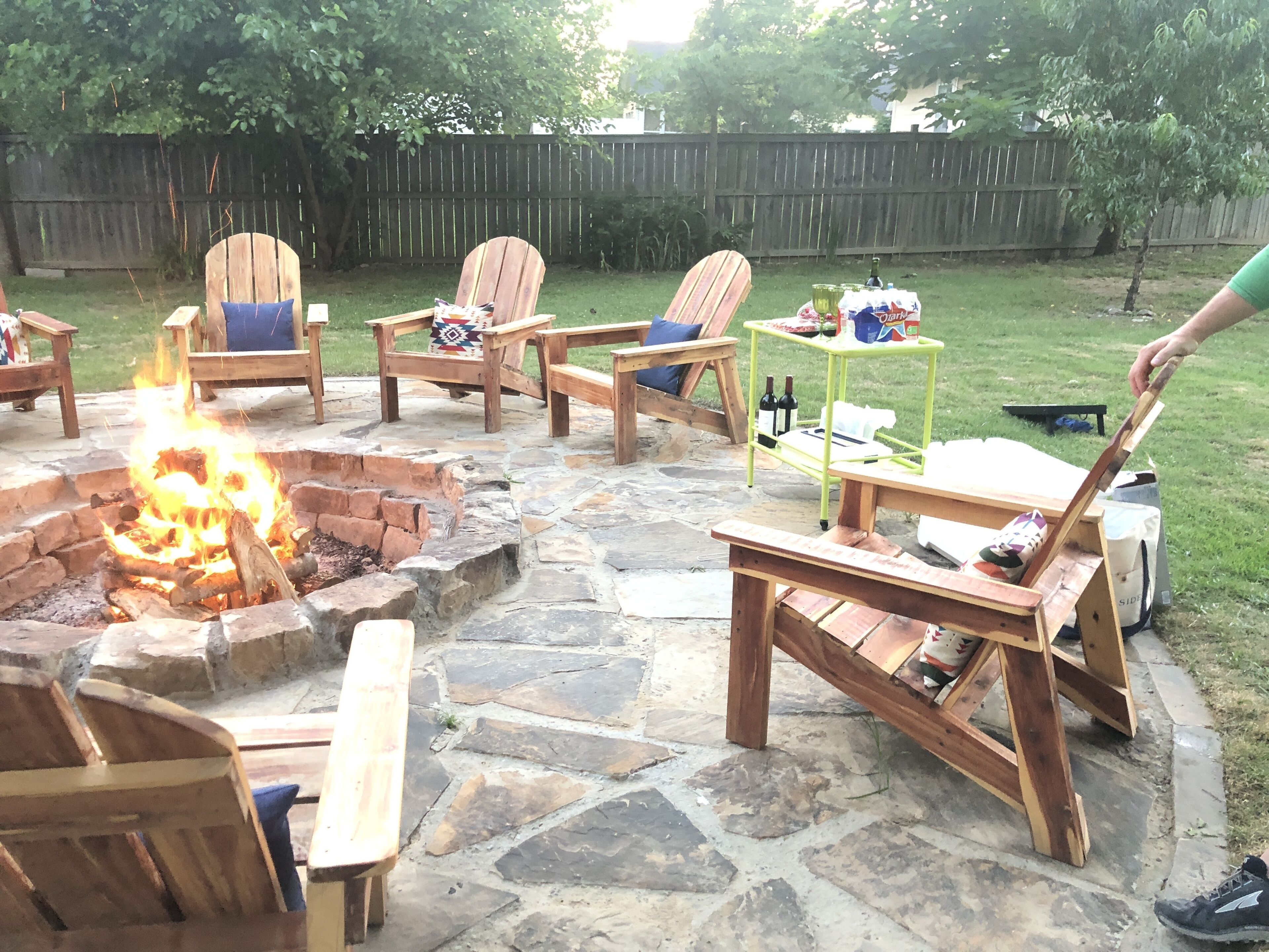 An outdoor patio area with a stone fire pit, wooden chairs, and a table.