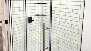 Eine Duschkabine mit Glastüren, einer weißen Fliesenwand, Ablage aus Glas und einem Metall-Duschkopf.