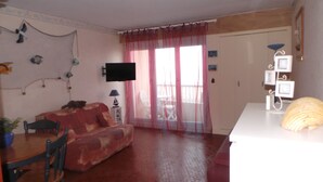 Un salon comprenant un canapé, un téléviseur fixé au mur, une table à manger avec des chaises et une fenêtre avec des rideaux.