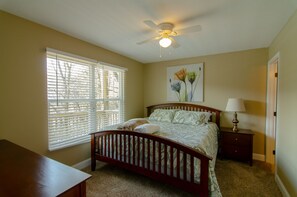 A bedroom with a bed, a ceiling fan, a nightstand with a lamp, and a window with blinds.