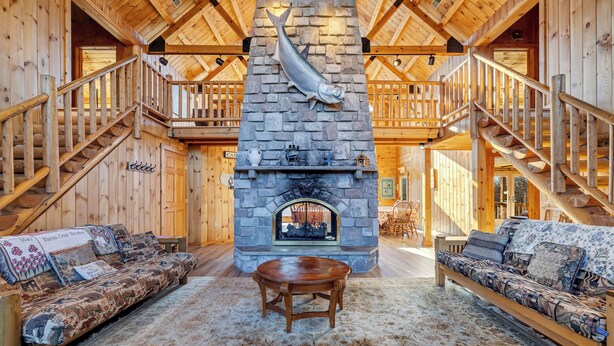 Ein Blockhaushaus-Interieur mit Kamin aus Stein, einer Holztreppe und zwei Sofas mit gemusterten Kissen.