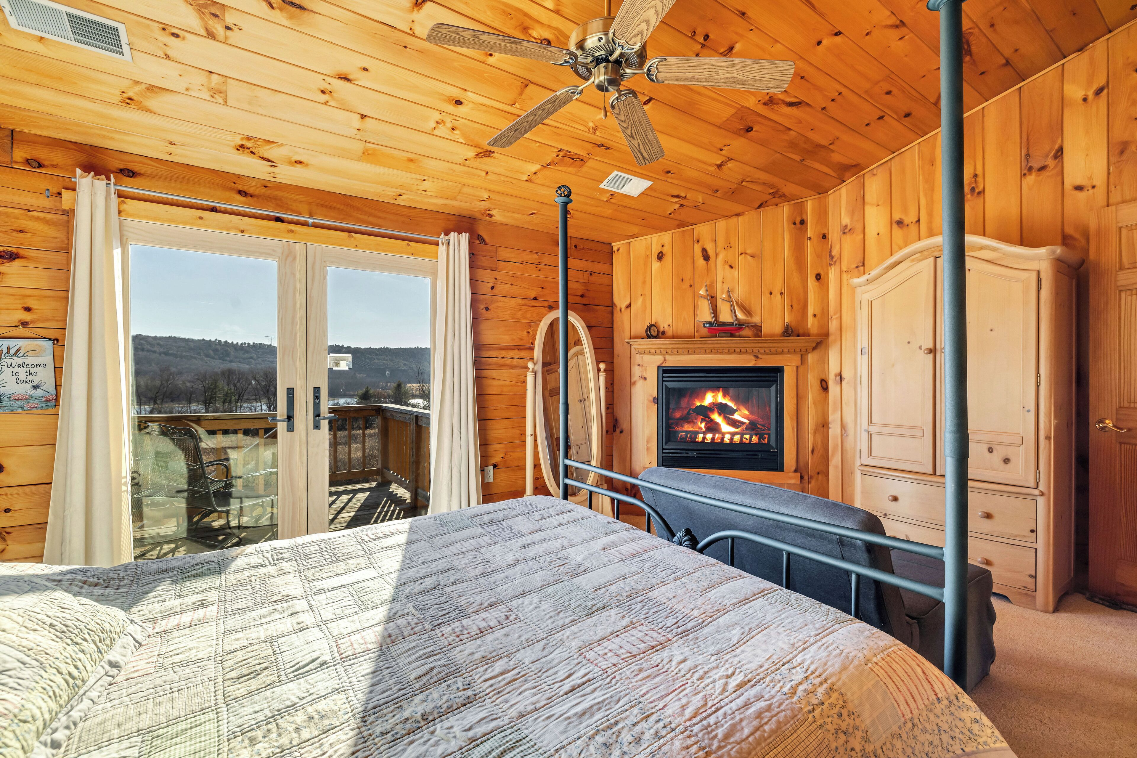 A wooden cabin bedroom with a bed, a fireplace, a ceiling fan, and a view of the outside.