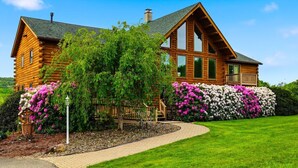 Eine Blockhütte mit breiter Veranda, einem Schornstein und einem gepflegten Garten mit blühenden Sträuchern.