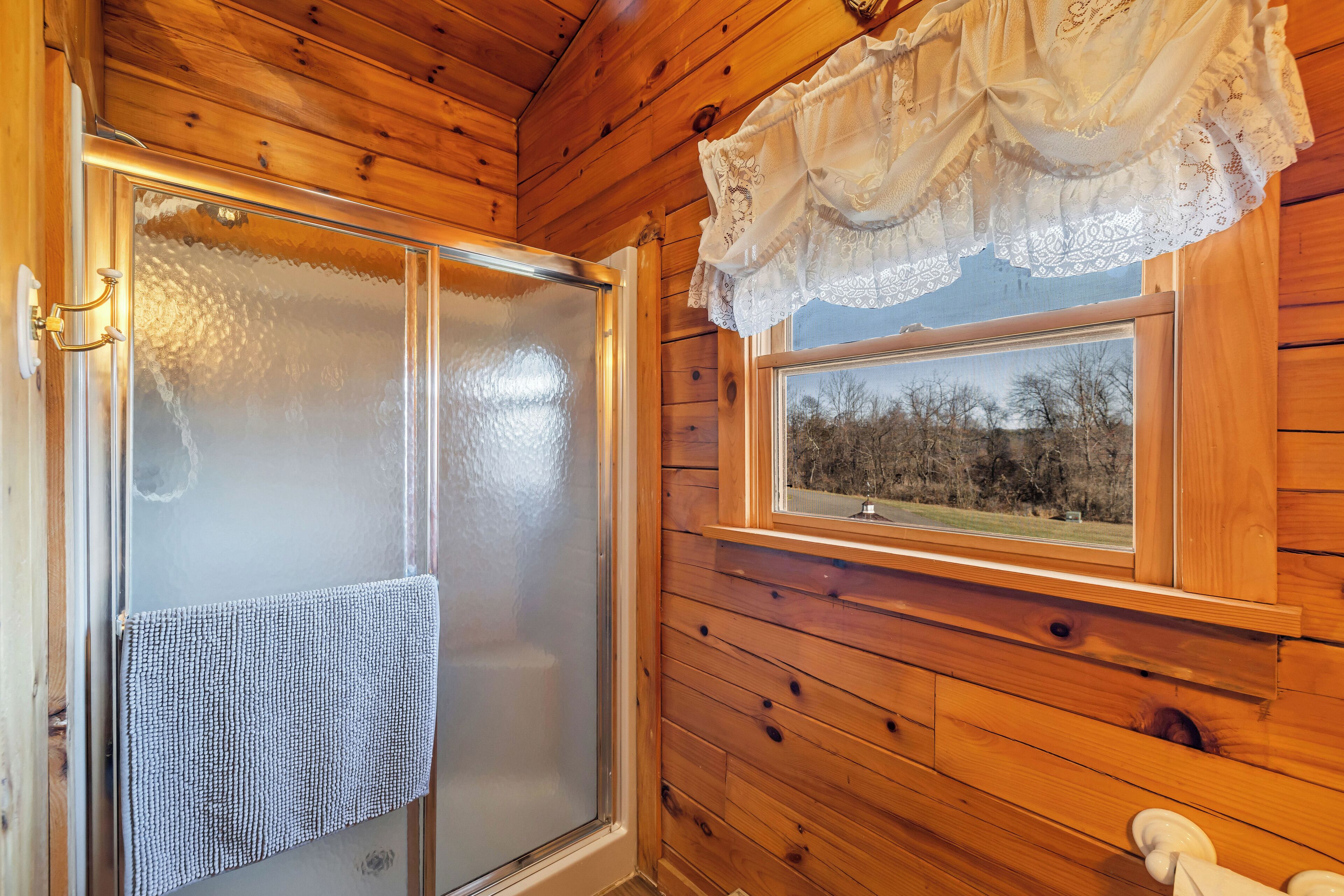 Interior de una cabaña de madera con una puerta de cristal empañado, una ventana con vistas a árboles y un encaje de cortina.