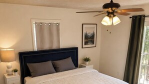 Ein Schlafzimmer mit einem Bett, einem Nachttisch, einer Lampe, einem Deckenventilator, einem Fenster mit Vorhängen und einem gerahmten Bild an der Wand.