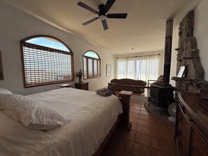 A bedroom with a bed, a ceiling fan, a stone fireplace, and large windows.