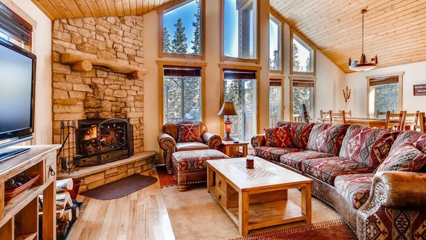 A cozy living room with a stone fireplace, wooden furniture, and large windows offering views of trees.