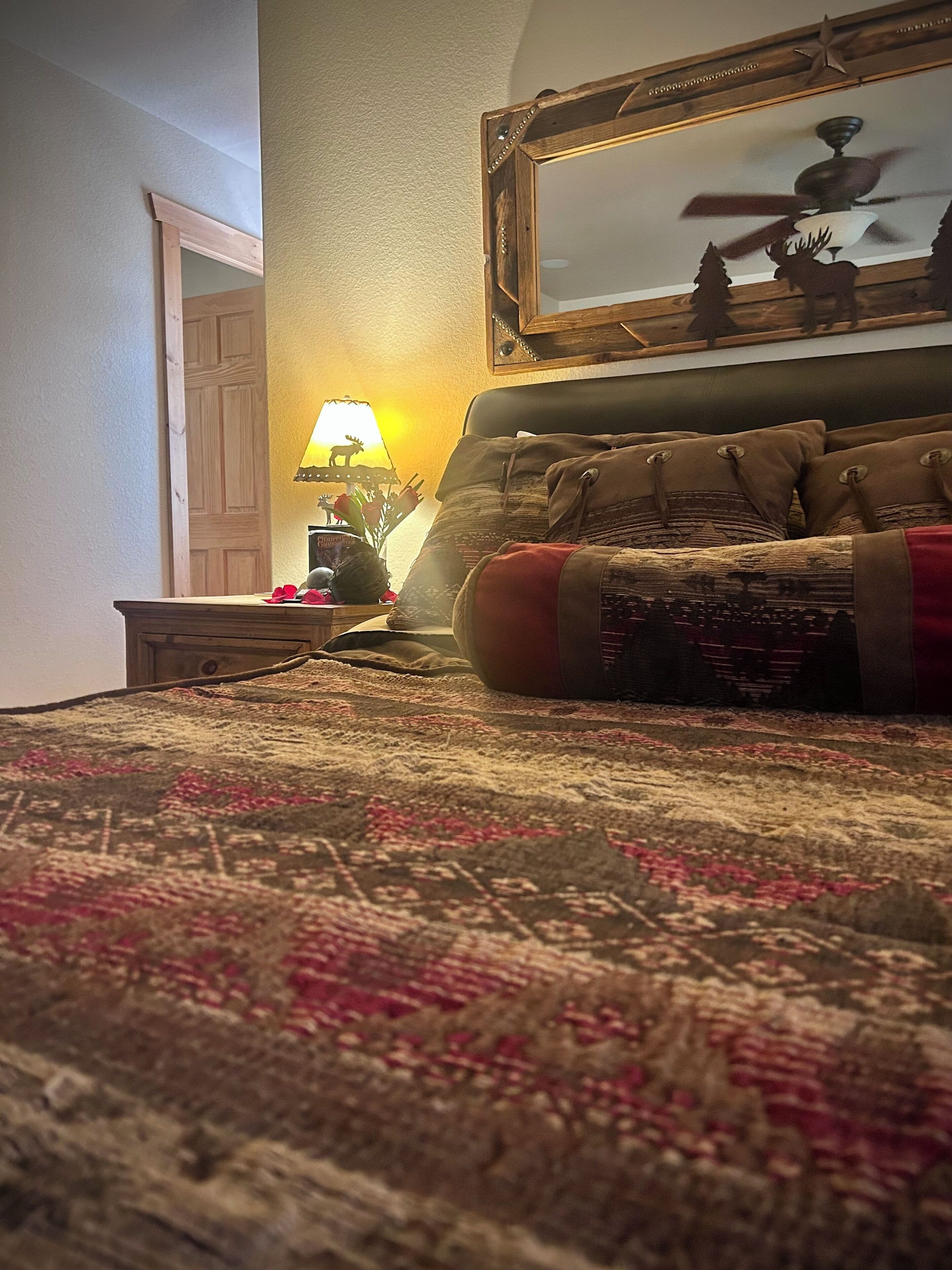 A bedroom with a patterned carpet, a bed with a red accent pillow, a nightstand with a lamp, and a mirror on the wall.
