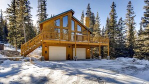 A two-story wooden cabin with a balcony, surrounded by snow and pine trees.