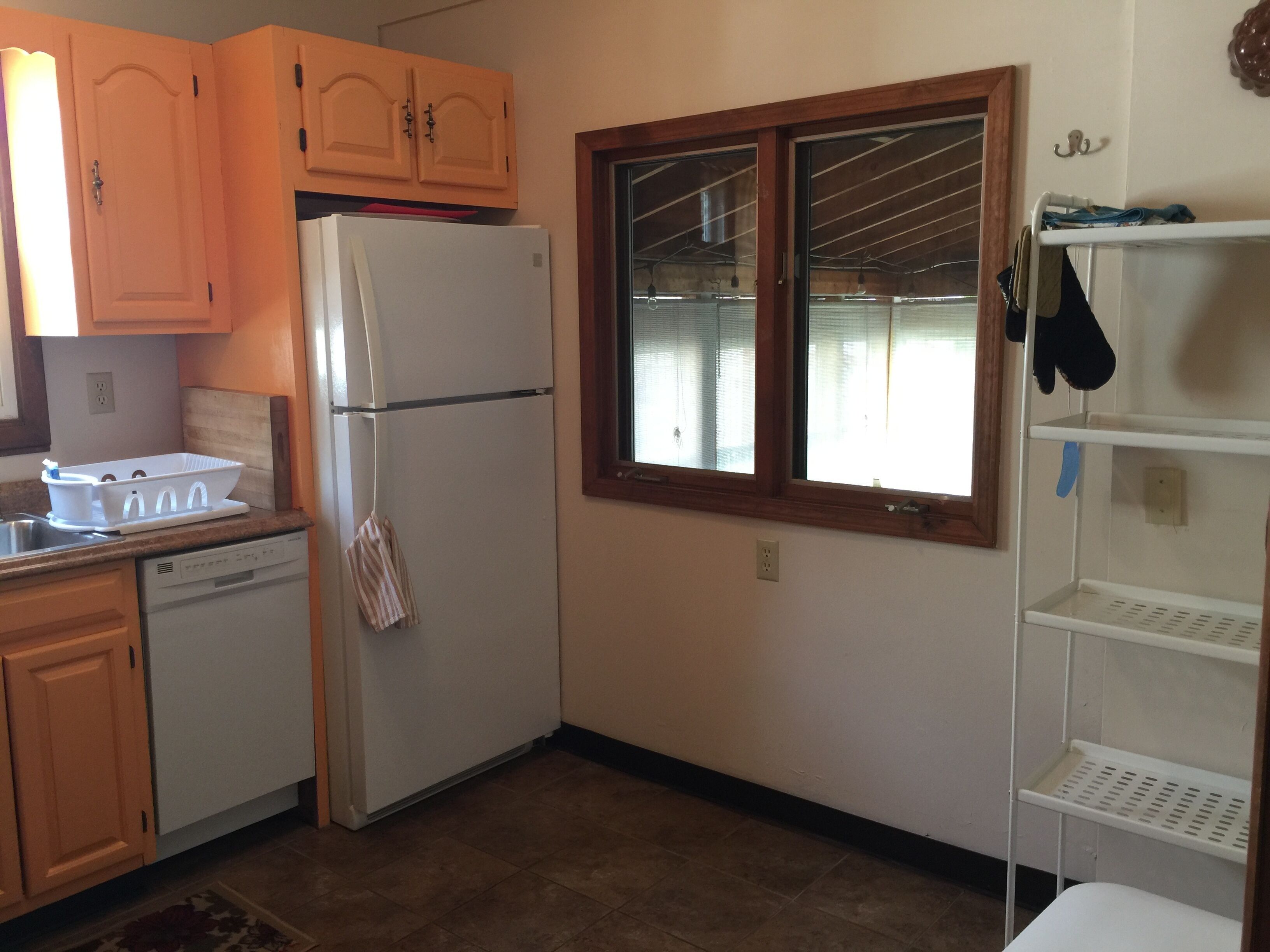 A kitchen with wooden cabinets, a white refrigerator, a window with blinds, and a white shelf unit.