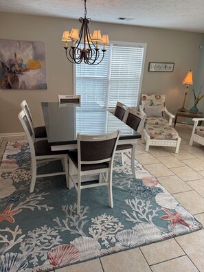 Un coin repas avec une table à plateau en verre, des chaises blanches, un lustre et un tapis à motifs floraux.