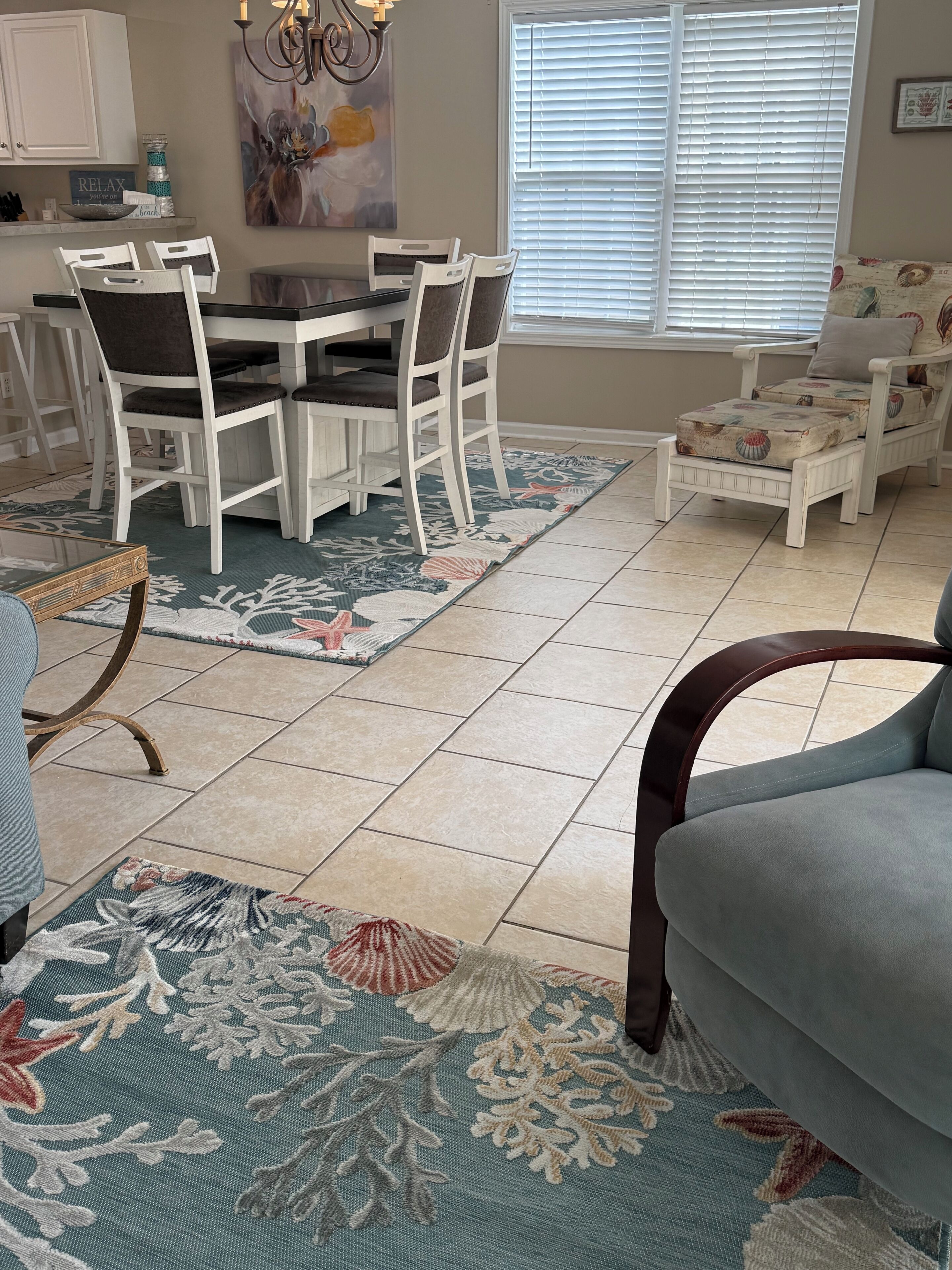 Un coin repas avec une table et des chaises blanches, une cuisine en arrière-plan, un tapis à motifs et une fenêtre avec des stores.