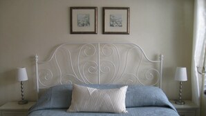Una cama con cabecera blanca, dos mesitas de noche con lámparas, y dos cuadros en la pared.