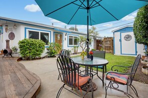 A patio with a table and chairs, a blue umbrella, and a house with blue trim and a white door. - Charming Vacation Flat: 5-minute walk to beach! (San Diego)