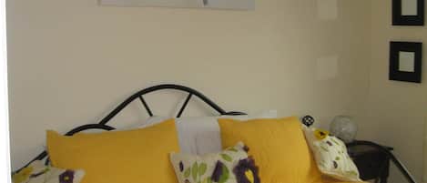 Un lit recouvert d’une couvre-lit jaune et agrémenté de plusieurs coussins, un mur orné d’un tableau représentant des oiseaux, et une petite table sur laquelle est posé un vase.
