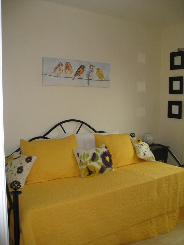 A bed with a yellow bedspread and multiple pillows, a wall with a painting of birds, and a small table with a vase. - Charming Vacation Flat: 5-minute walk to beach! (San Diego)