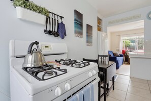 Een keuken met een wit fornuis, een waterkoker en een klok. Er staat een eettafel met stoelen en er is een blauwe bank op de achtergrond.