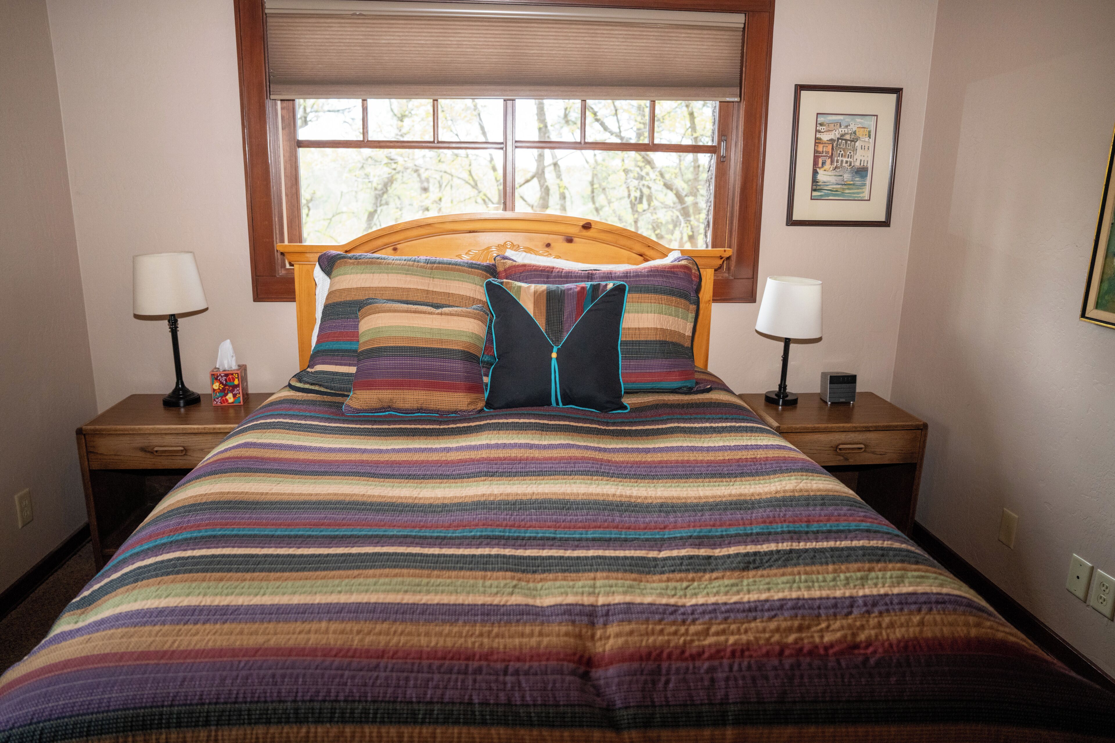 Una cama con colcha a rayas, dos mesitas de noche con lámparas, una ventana con persianas y un cuadro en la pared.
