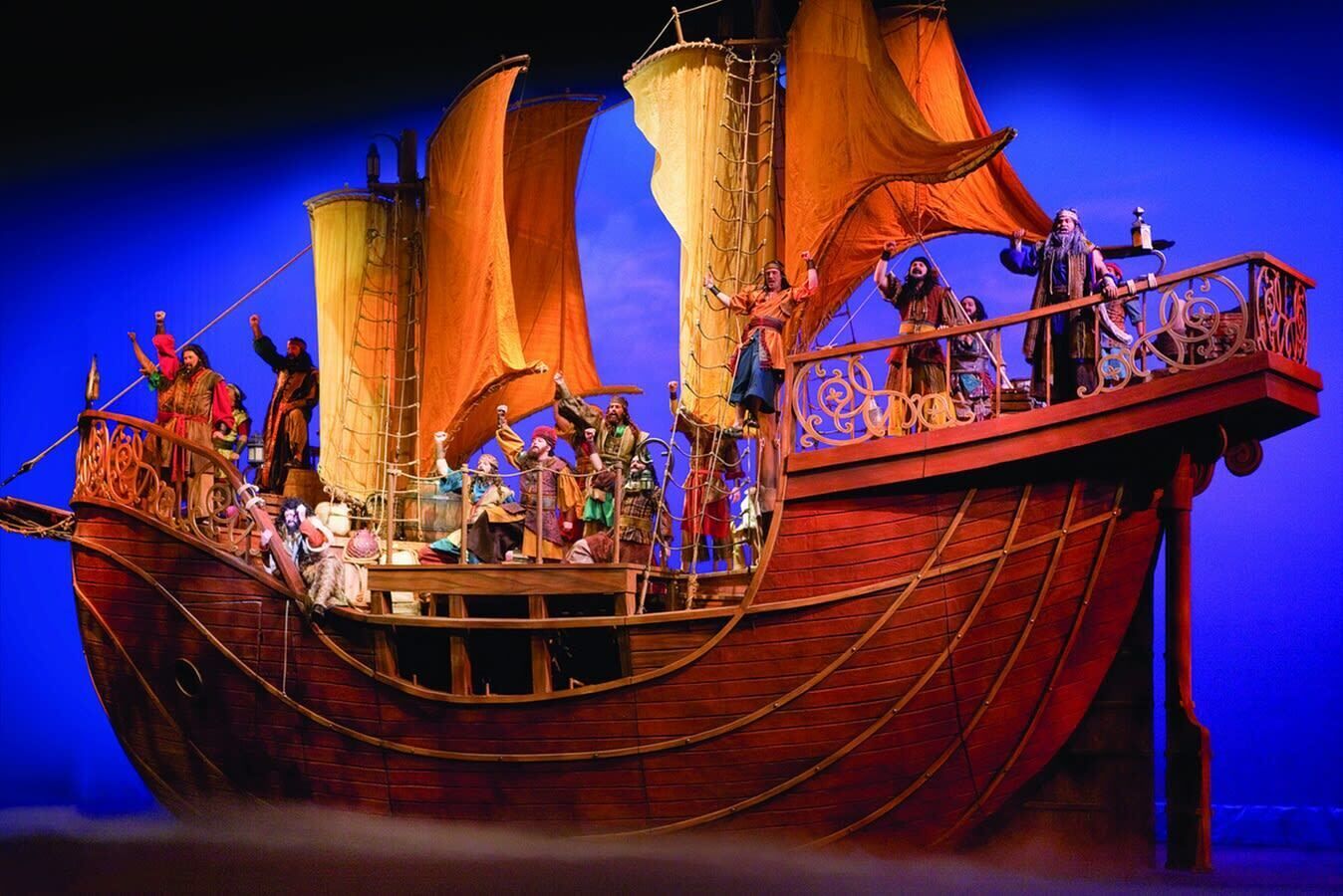 A stage with a large ship set, featuring multiple characters in period costumes, a wooden ship deck, and a blue background.