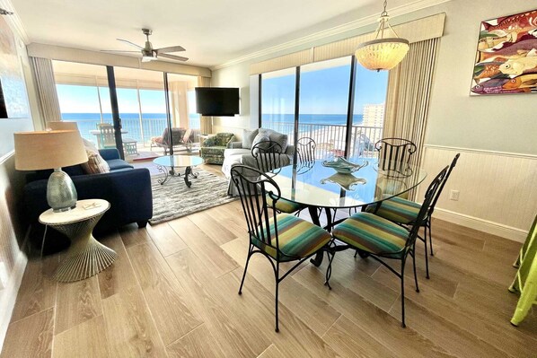 A spacious living area with a balcony, a dining table with chairs, and a television mounted on the wall. - 3BR Beachfront Gem | Huge Balcony + Stunning Views | Nearly $100K Reno! (Orange Beach)