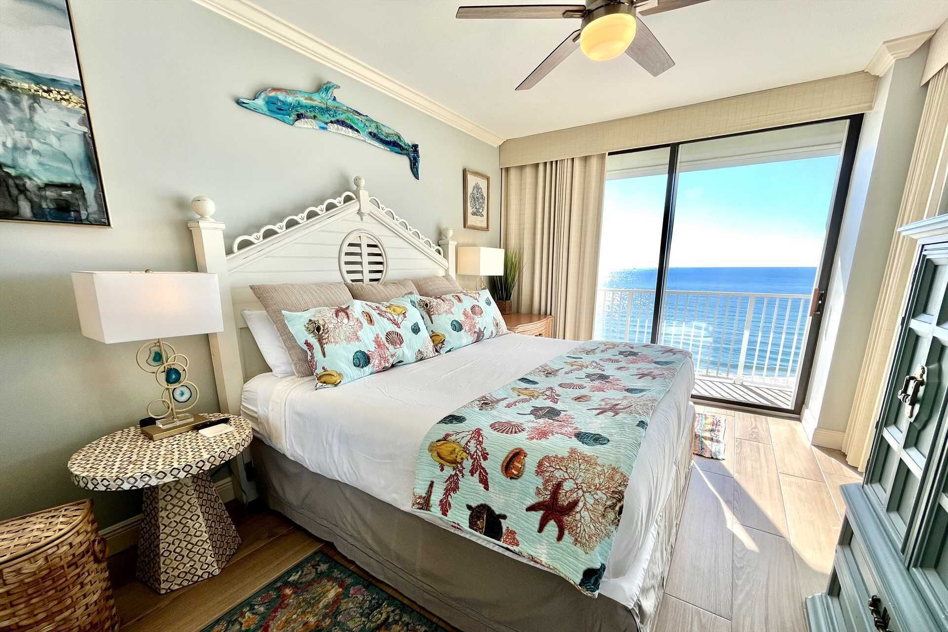 Un dormitorio con cama, mesita de noche, lámpara, ventilador de techo, ventana con vista al océano y una pieza decorativa de delfín en la pared.