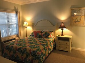Un dormitorio con cama, dos lámparas de noche, una mesita de noche, una ventana con persianas y un cuadro enmarcado en la pared.