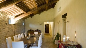 A dining area with a long wooden table, white upholstered chairs, and a patterned tablecloth.