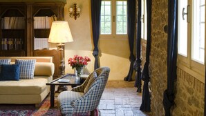 A cozy room with a stone wall, a wooden ceiling, a patterned rug, a sofa, a chair, a lamp, and a bookshelf.