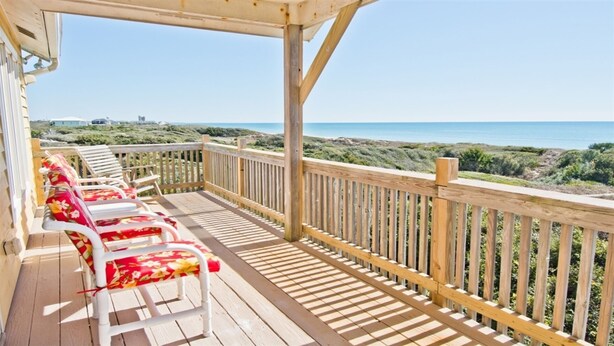 Terraza de madera con sillas blancas y barandilla de madera que da a una playa.