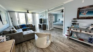 A spacious living room with a grey sofa, a coffee table, and a large window. There is a door leading to another room with a bed and a painting on the wall. - Ocean Village Vacation Rental, Golf view. Short walking distance to the beach.  (Hutchinson Island)